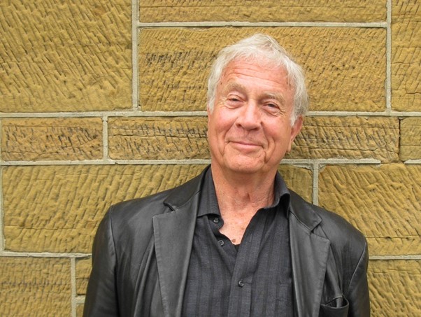 Professor Henry Reynolds, standing in front of an ochre-coloured brick wall. He has short grey hair and is wearing a black shirt and jacket, and is smiling at the camera. 